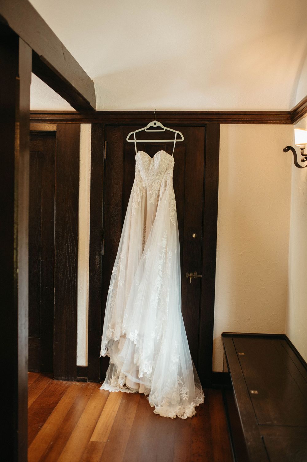 Lace wedding dress hanging on white hanger against dark wood trim interior.