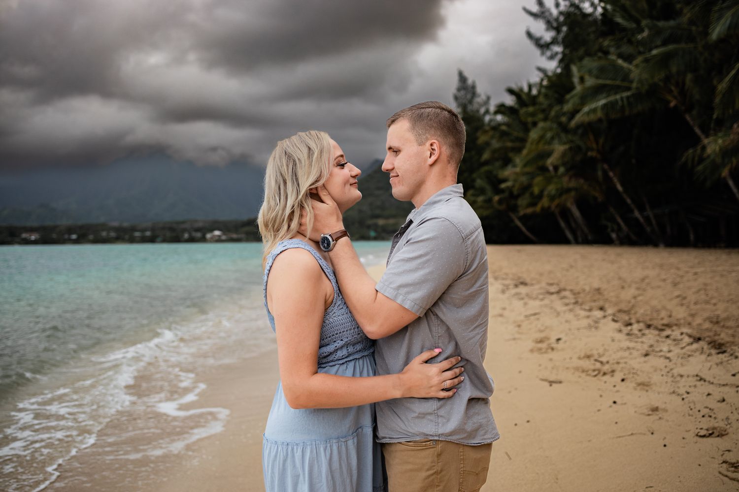 Romantic couple embraces on a tropical beach with stormy clouds overhead and turquoise waters in the background.