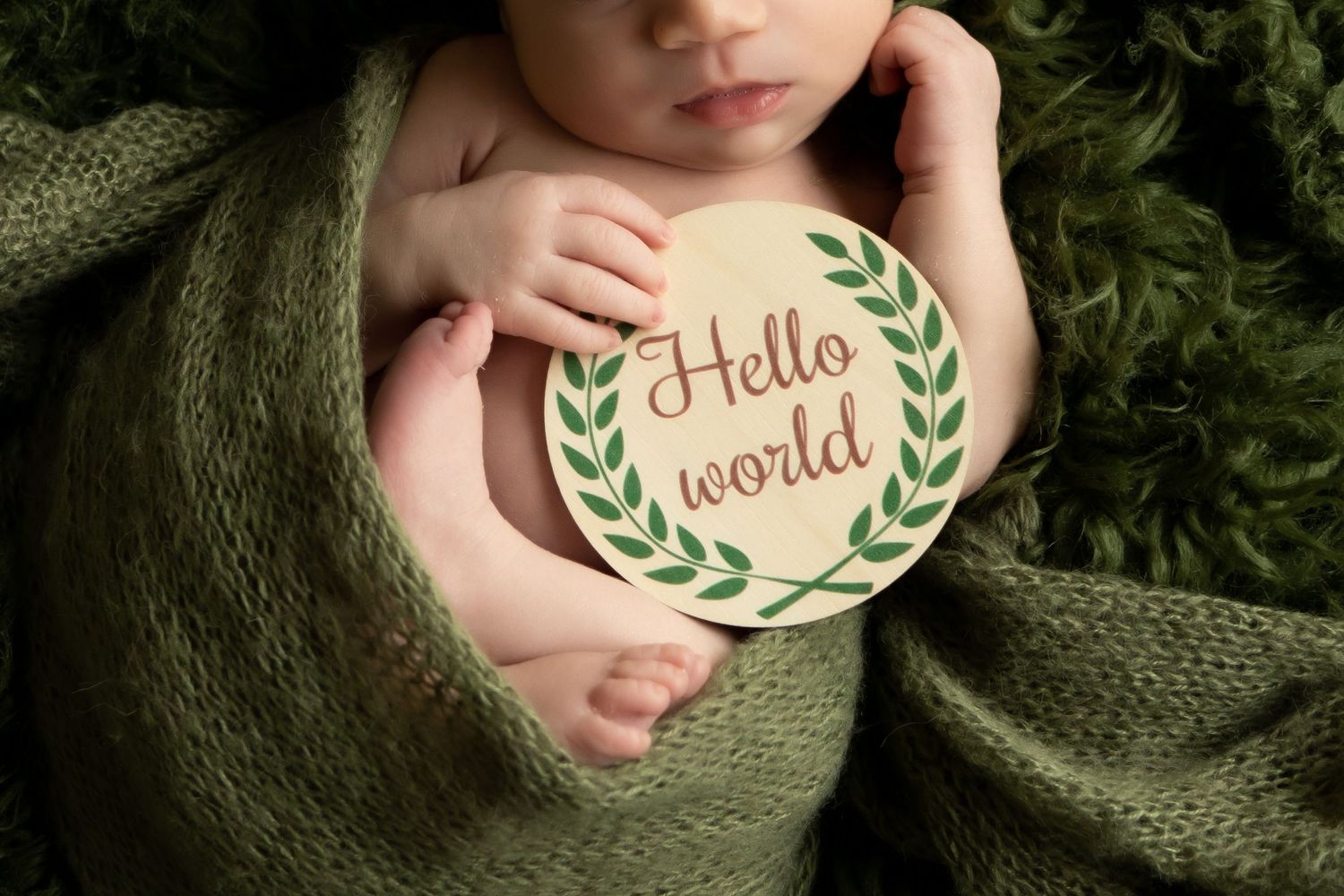 Newborn baby wrapped in green blanket holding Hello World wooden sign.