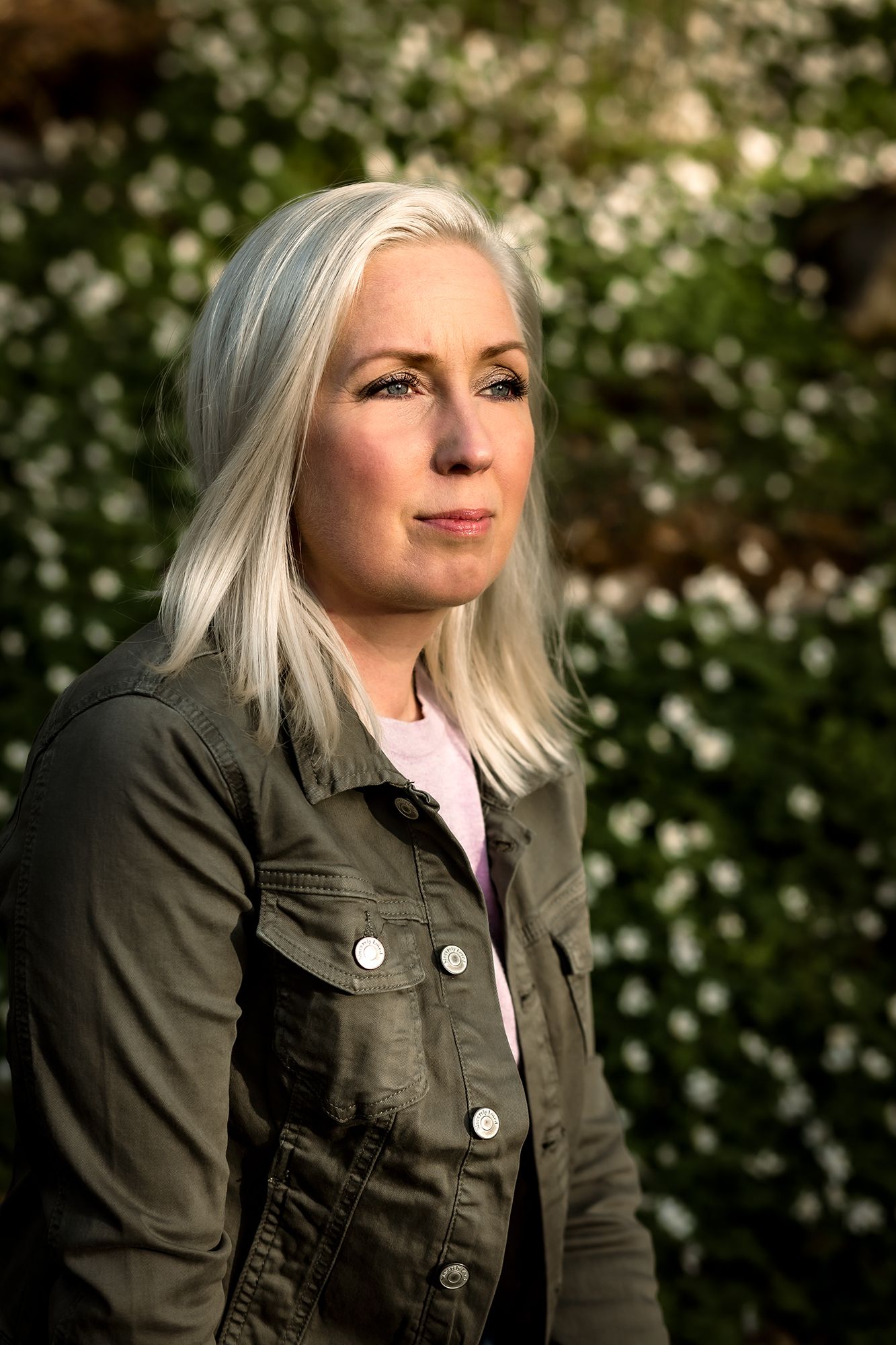 Portrait of a person in an olive jacket against flowering bushes at golden hour.