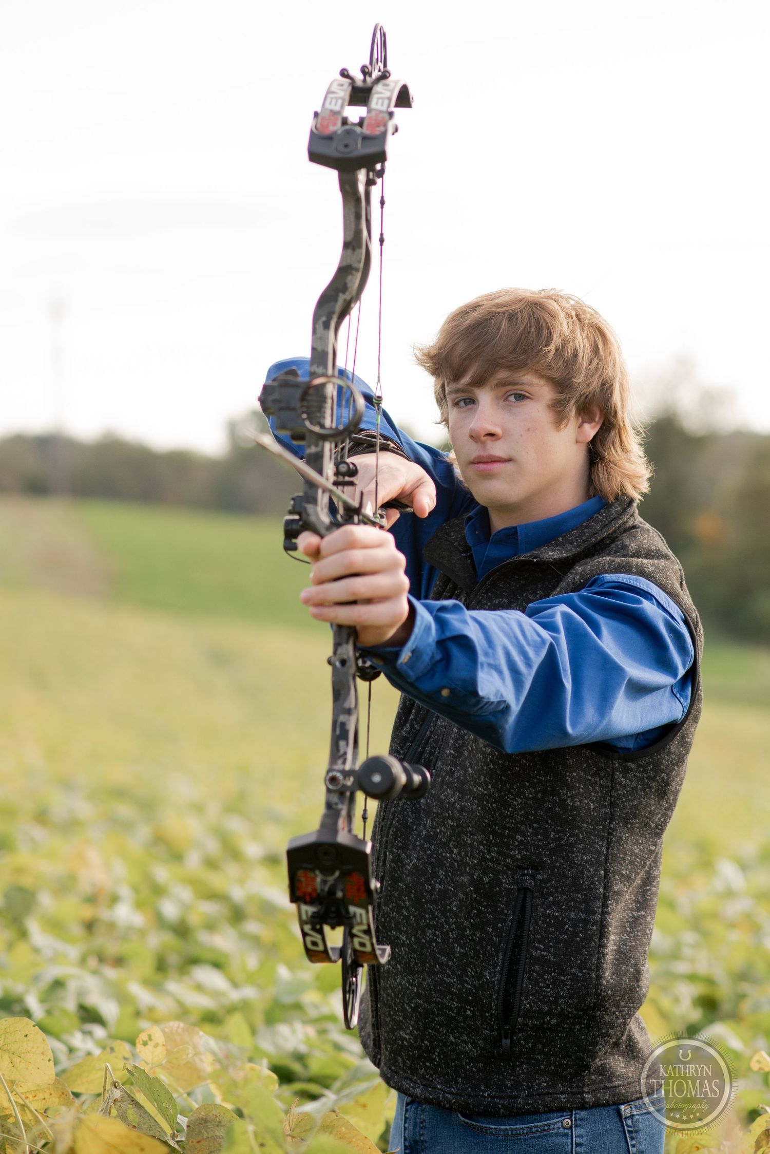 Someone aims a compound bow while wearing a blue jacket and gray vest in a field.