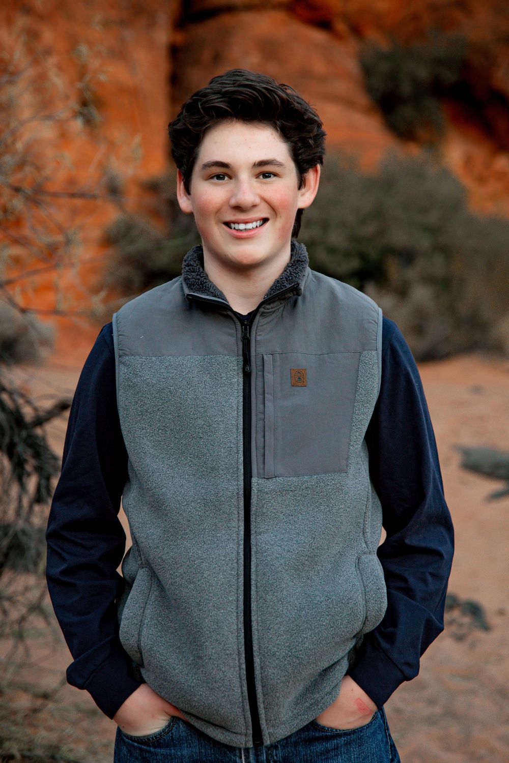 A teen in a gray fleece vest and navy sleeves stands in front of red rock formations.