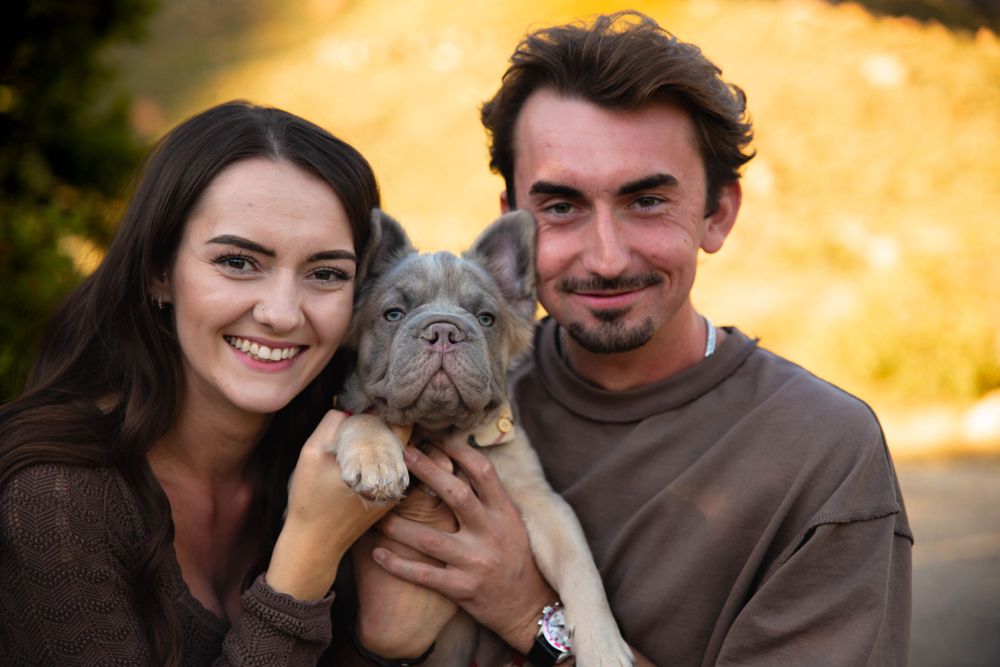 Couple holding their wrinkly French Bulldog puppy against golden sunset background.