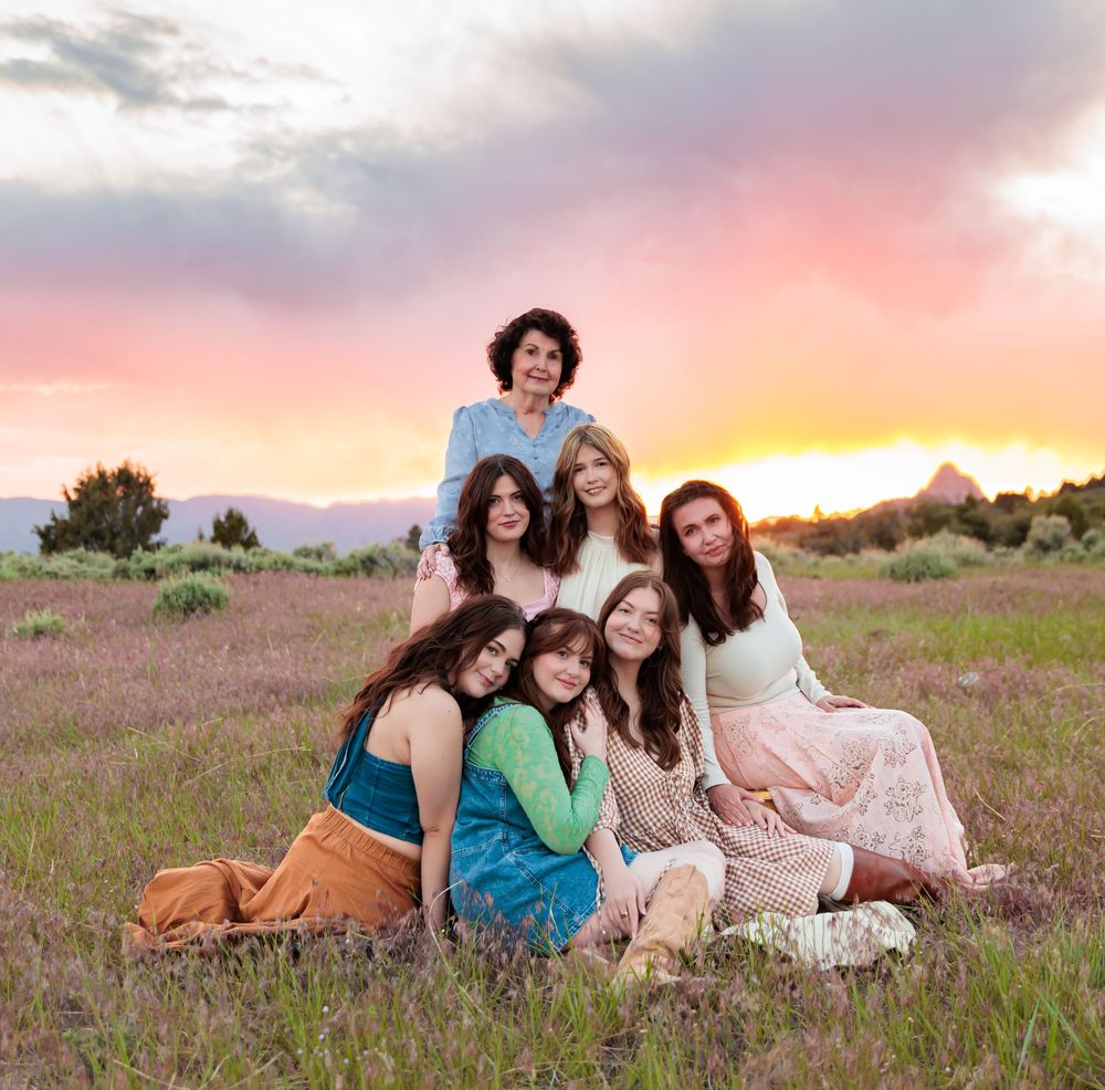 Group of seven women posing together in a field at sunset with colorful pink and orange sky backdrop.