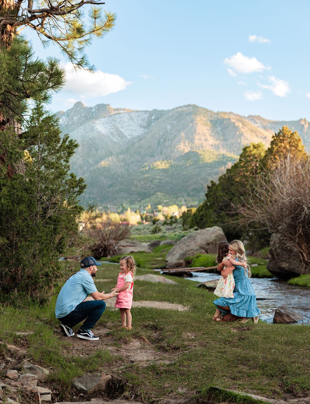 Family enjoying a scenic mountain stream with pine trees and rocky peaks in the background on a sunny day.