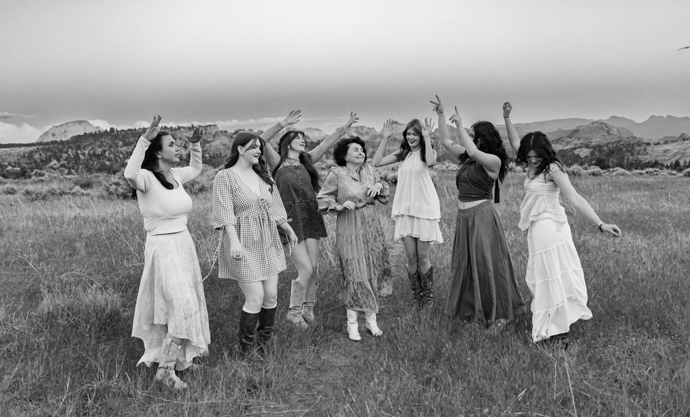 A black and white photo of women dancing joyfully in an open field with mountains in the distance.