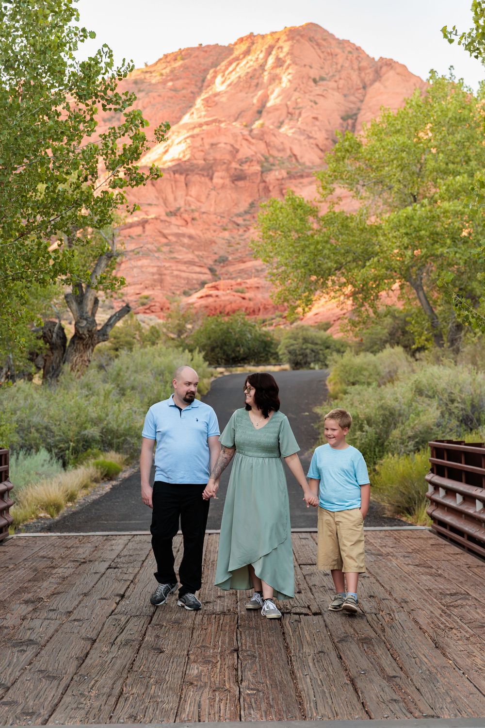 Family of three walking on a wooden bridge with red rock mountains and green trees in the background at sunset.