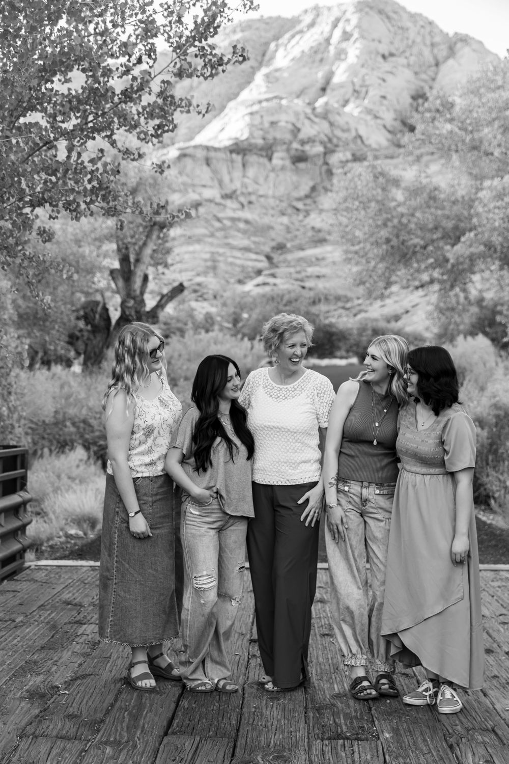 Five women laughing together on a wooden bridge with rocky mountain landscape in black and white photo.