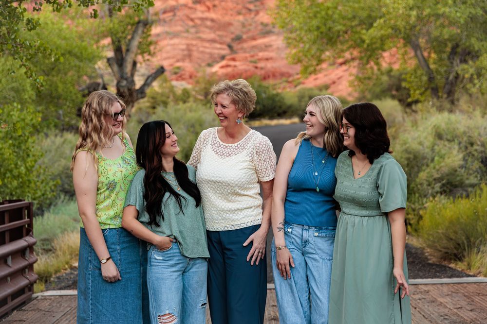 Five women smiling and laughing together on a bridge with red rock formations in the background.