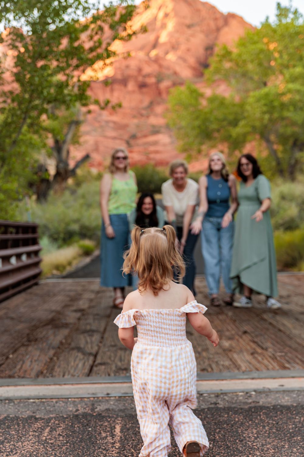 Toddler girl in pink romper running toward group of women on bridge with red rock mountains behind.
