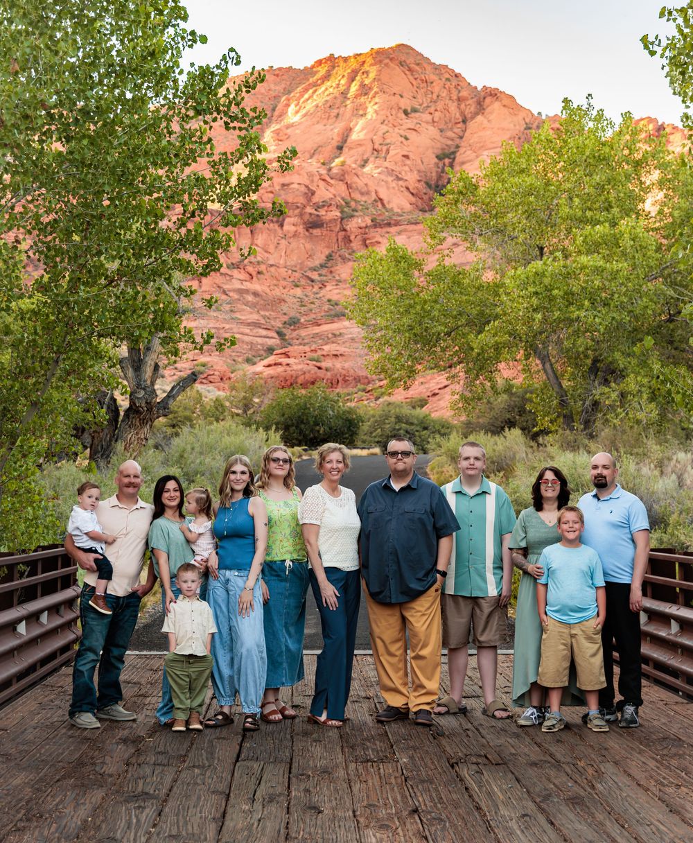 Large family group posing on a wooden bridge with red rock canyon cliffs and green trees in the background.
