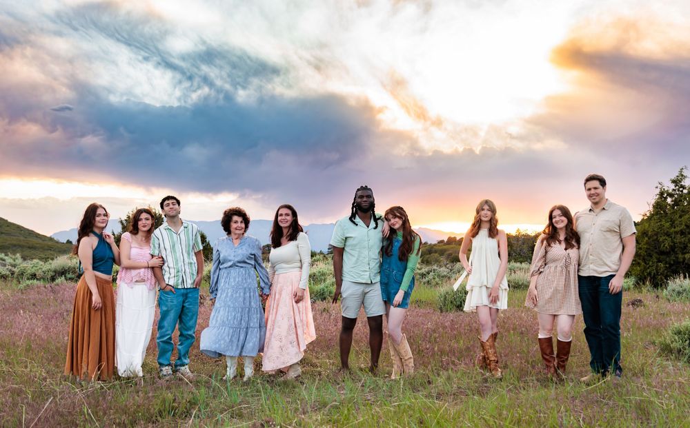 Group of nine people posing in a field at golden hour sunset wearing colorful casual outfits.