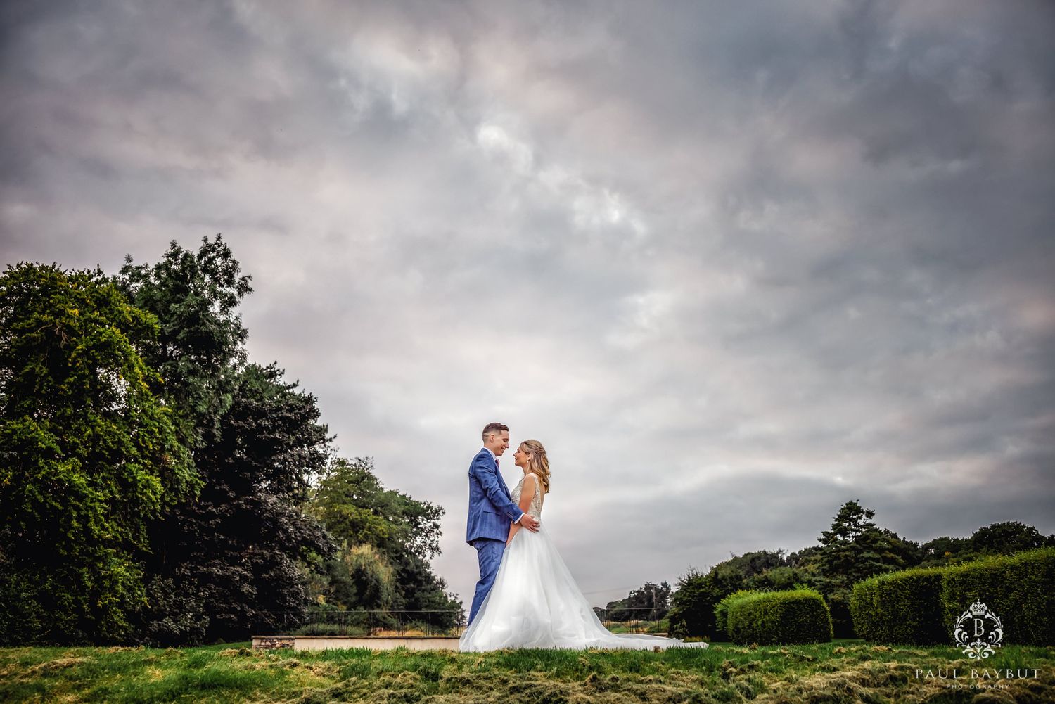 Romantic wedding moment captured against dramatic cloudy sky and lush green landscape.