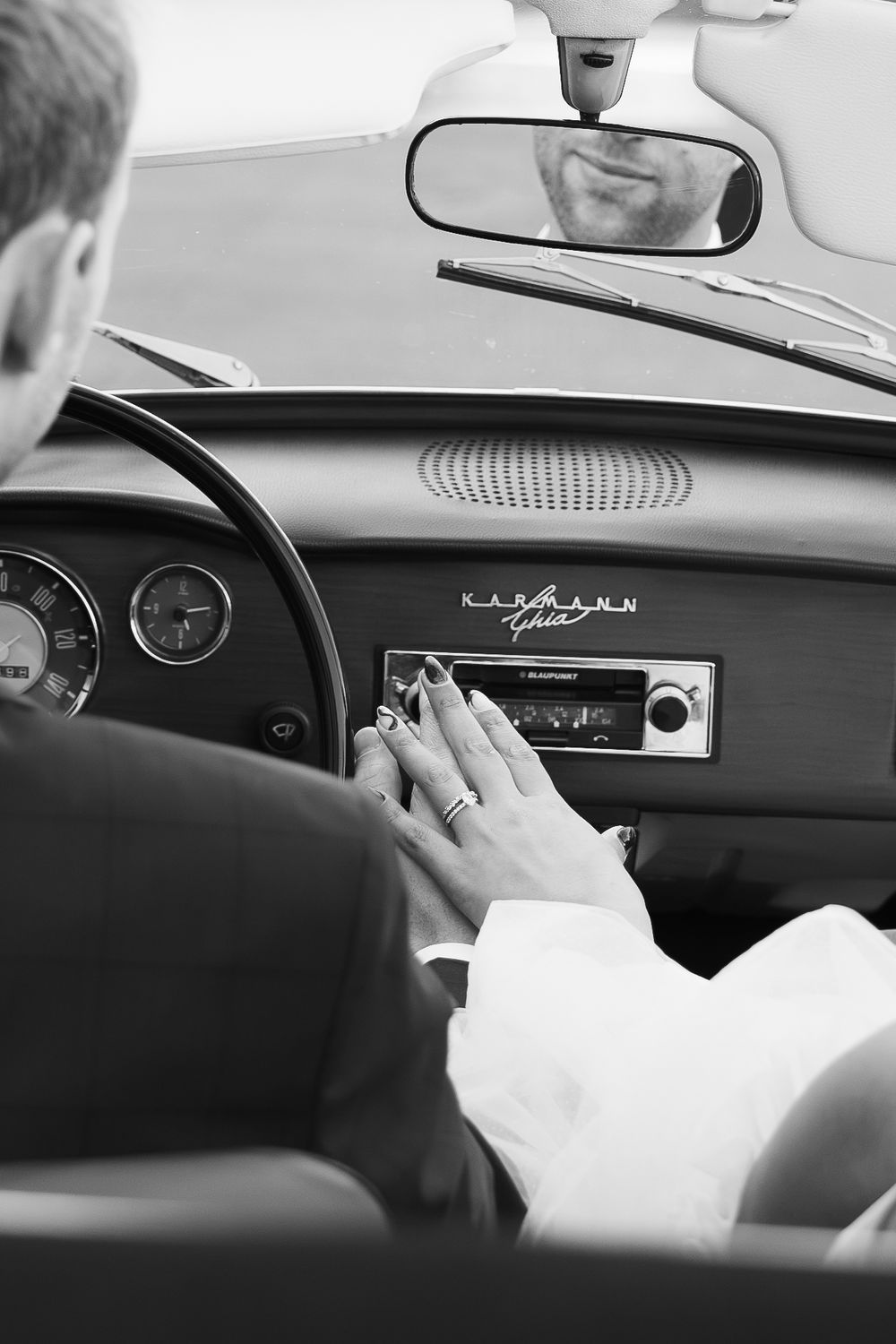 Zwart-witfoto van de handen van het bruidspaar in een witte oldtimer.