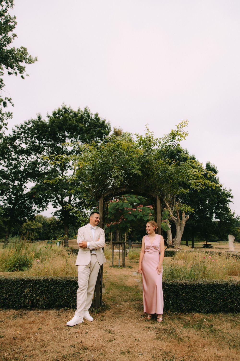 A couple in elegant attire stand in a lush garden, man in beige suit, woman in pink dress.
