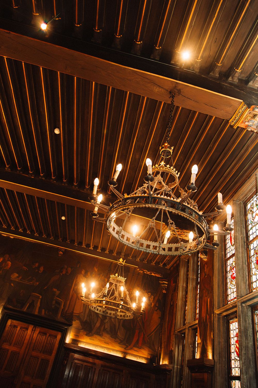 Gothic hall with ornate iron chandeliers, wooden beam ceiling, stained glass windows, and warm candlelight ambiance.