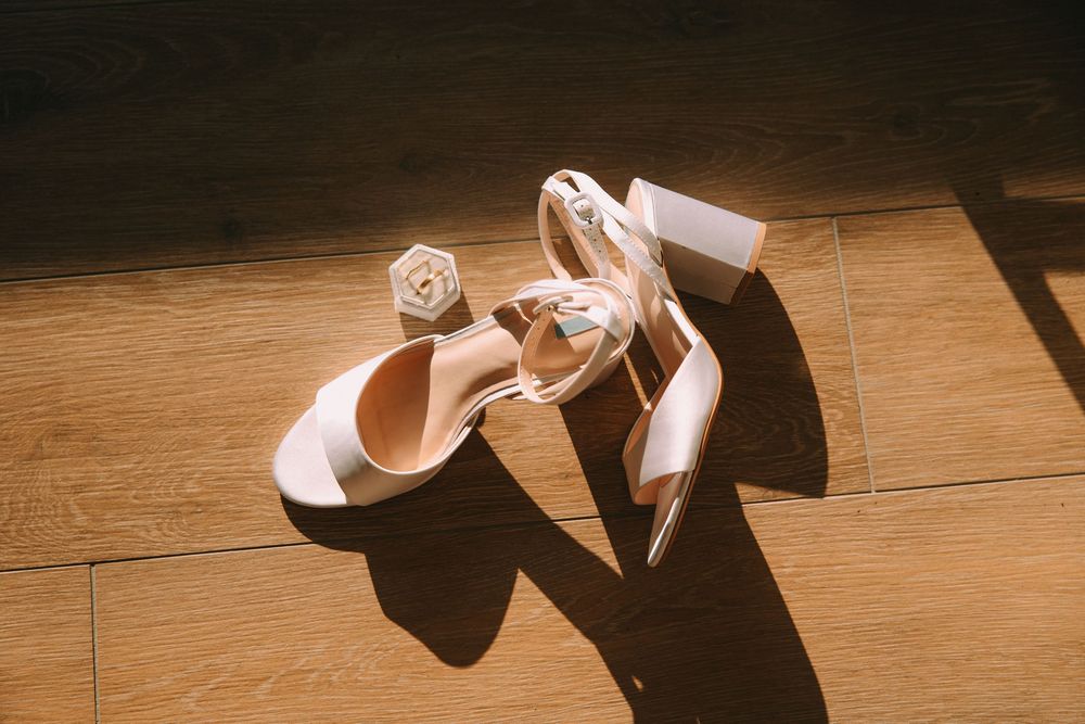 White block heel sandals with ankle strap on wooden floor in sunlight, with small floral accessory.