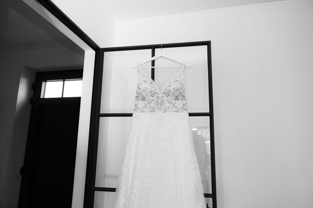 Elegant lace wedding dress hanging on a black frame rack in a bright room, captured in black and white photography.