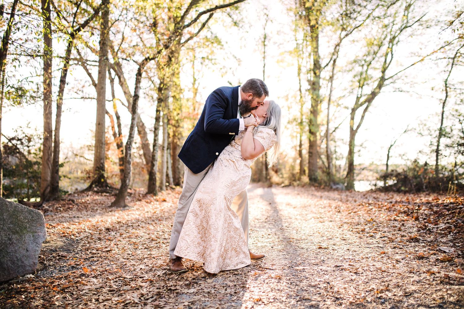 Couple sharing romantic dip kiss on autumn leaf-covered path surrounded by trees.