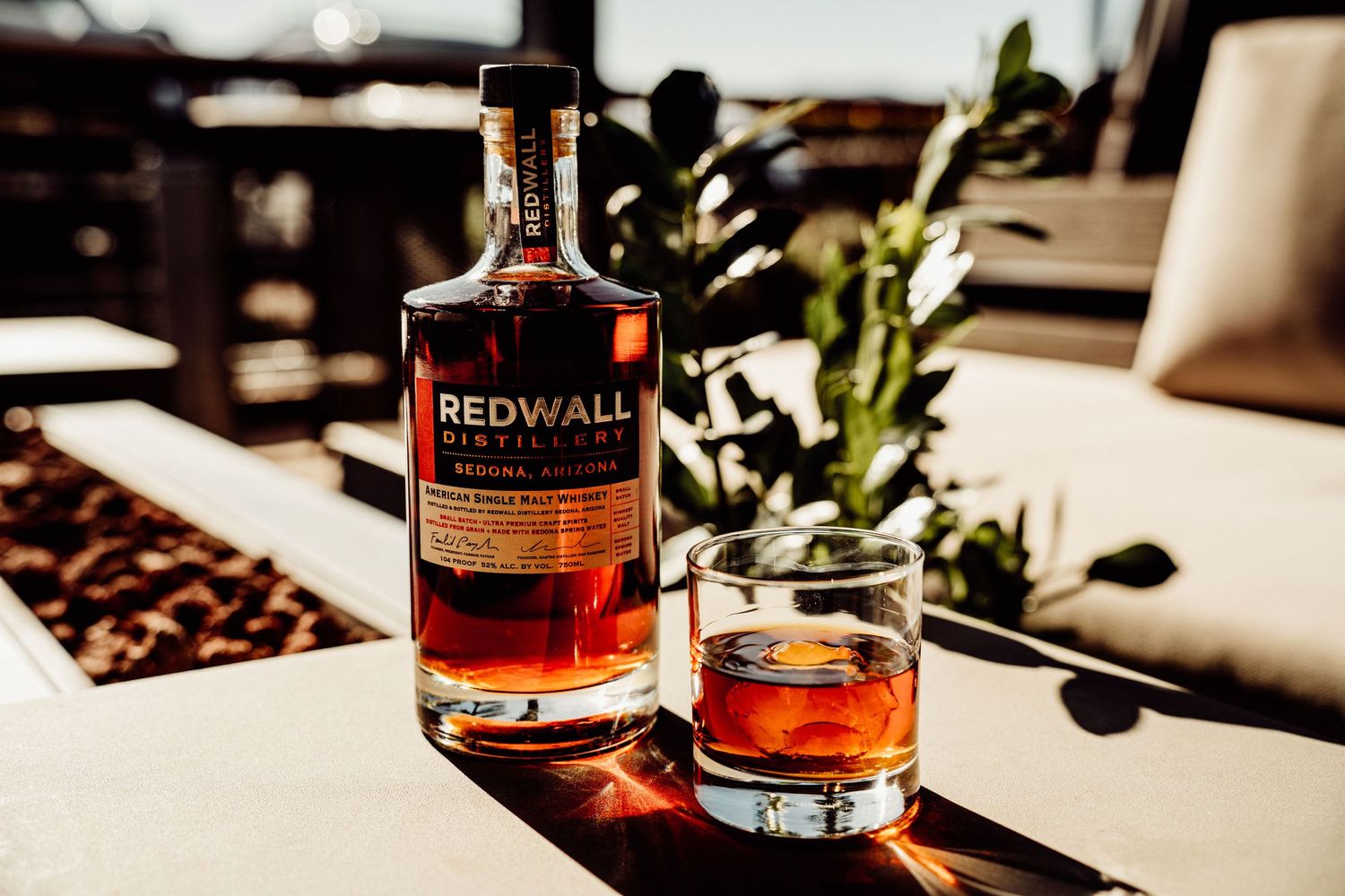 Redwall whiskey bottle and glass on table with warm lighting and decorative plants in background.