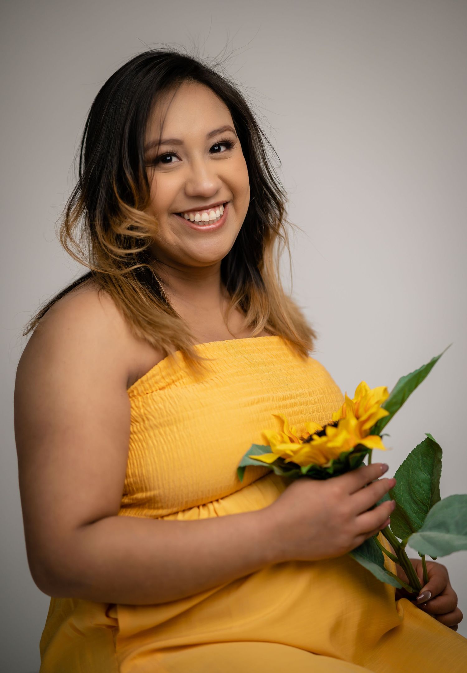 Joyful studio portraits with sunflower props and yellow dress against neutral backdrop.