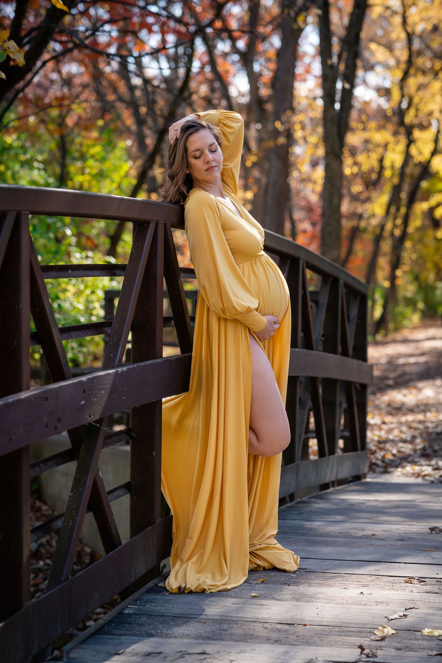 Maternity session on a wooden bridge surrounded by autumn foliage featuring a flowing yellow gown.
