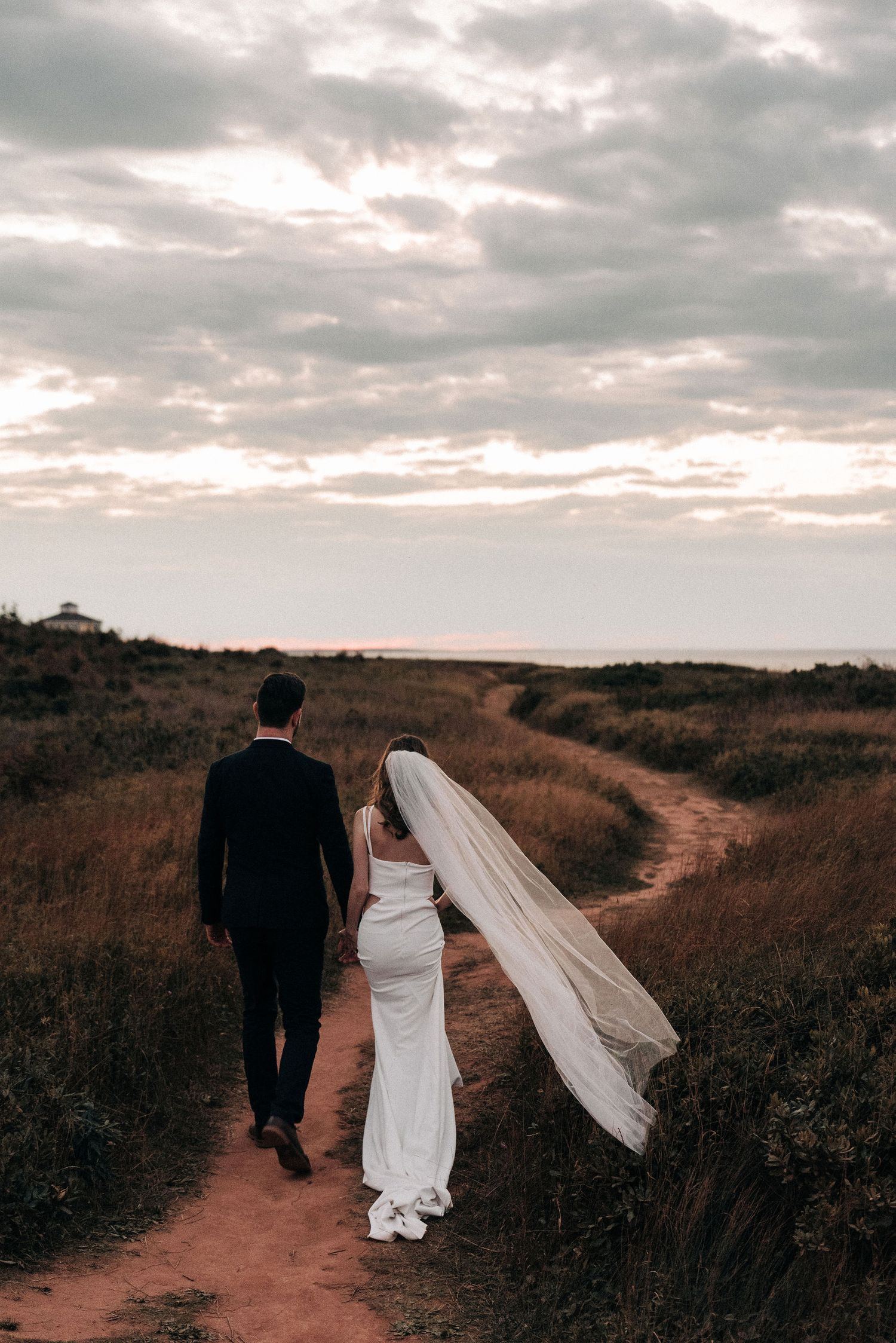 Wedding couple walking down dirt path with flowing veil and moody sky.
