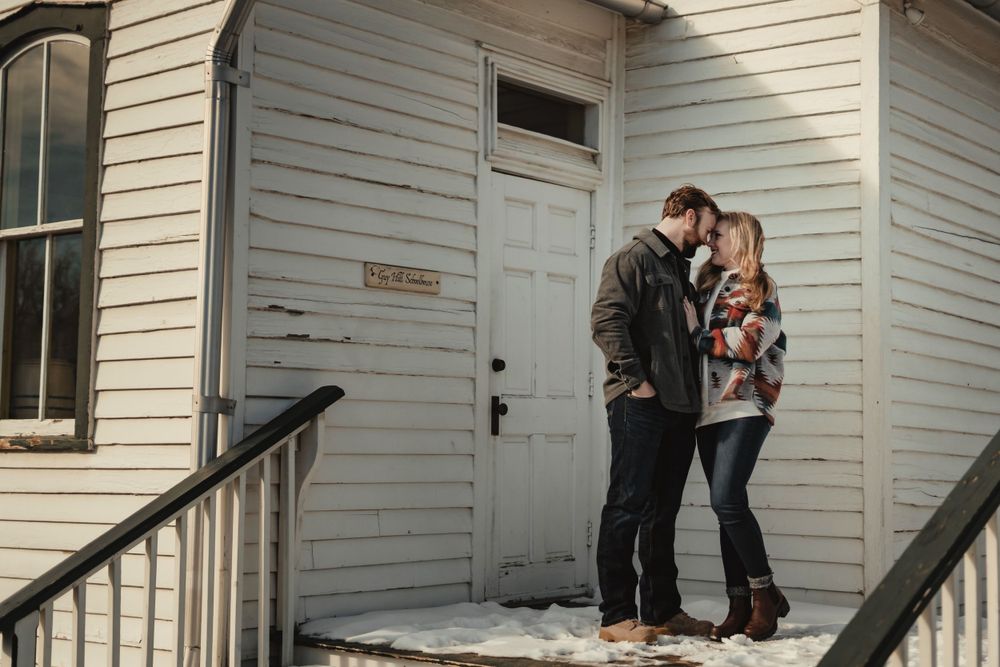 A couple stands intimately on a snow-dusted porch of a white wooden building.