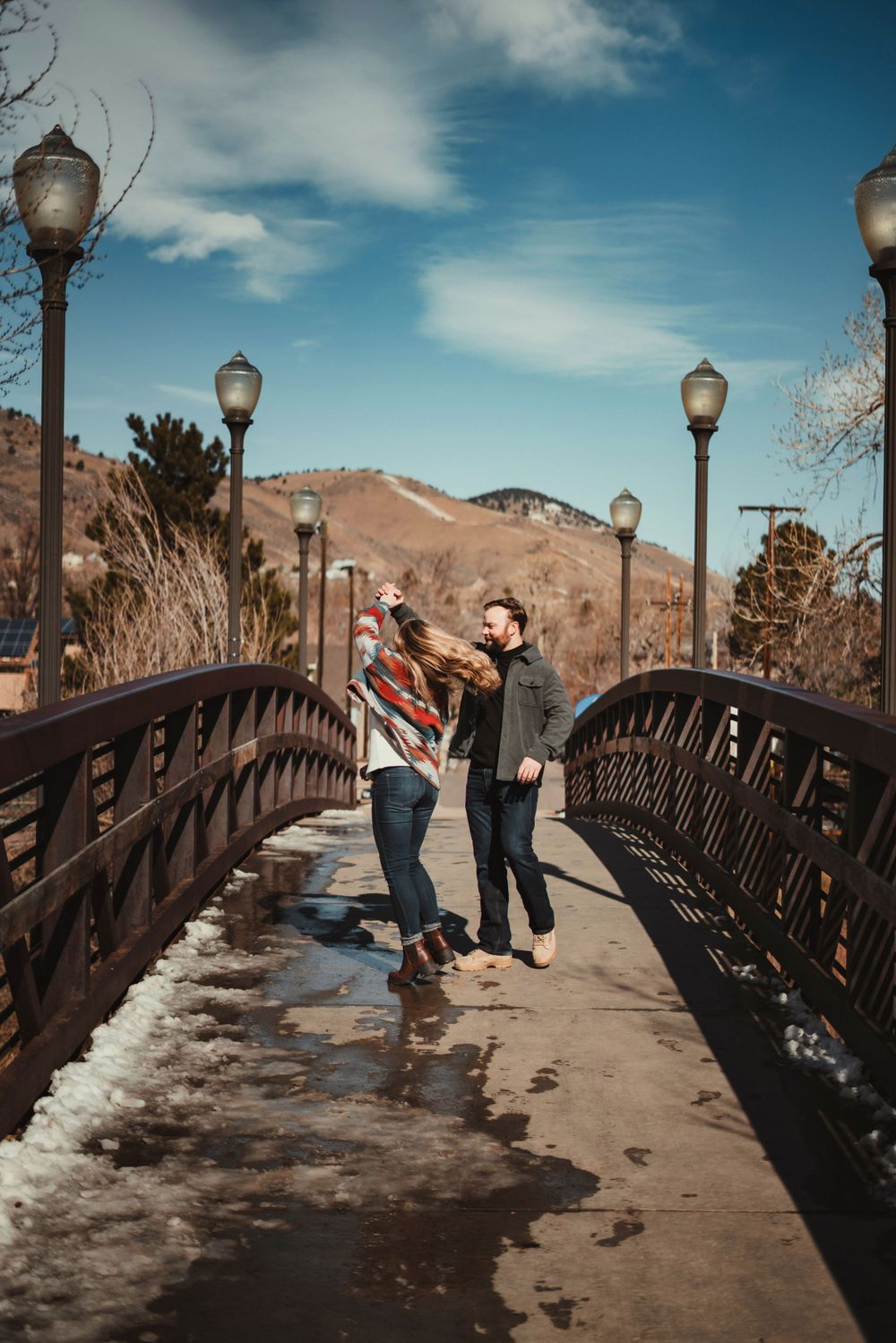 A couple shares a romantic moment on a bridge with mountain scenery and lamp posts.