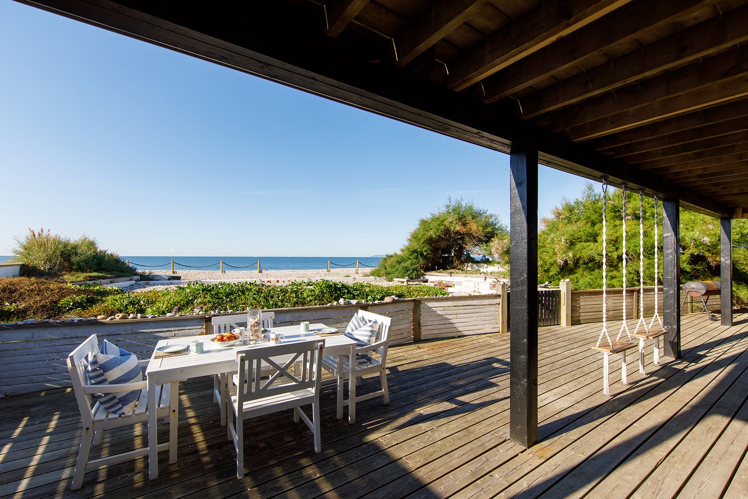 Oceanfront deck with white dining furniture and beautiful sea views through exposed wooden beams.