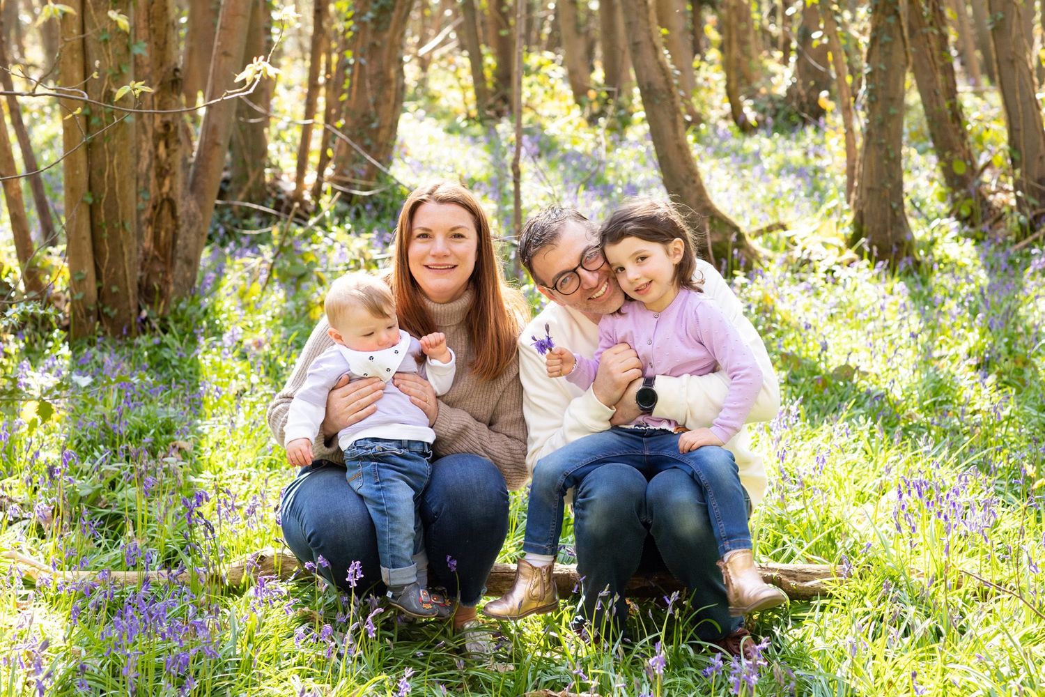 A family group sits together on grass surrounded by springtime bluebells in a wooded setting.