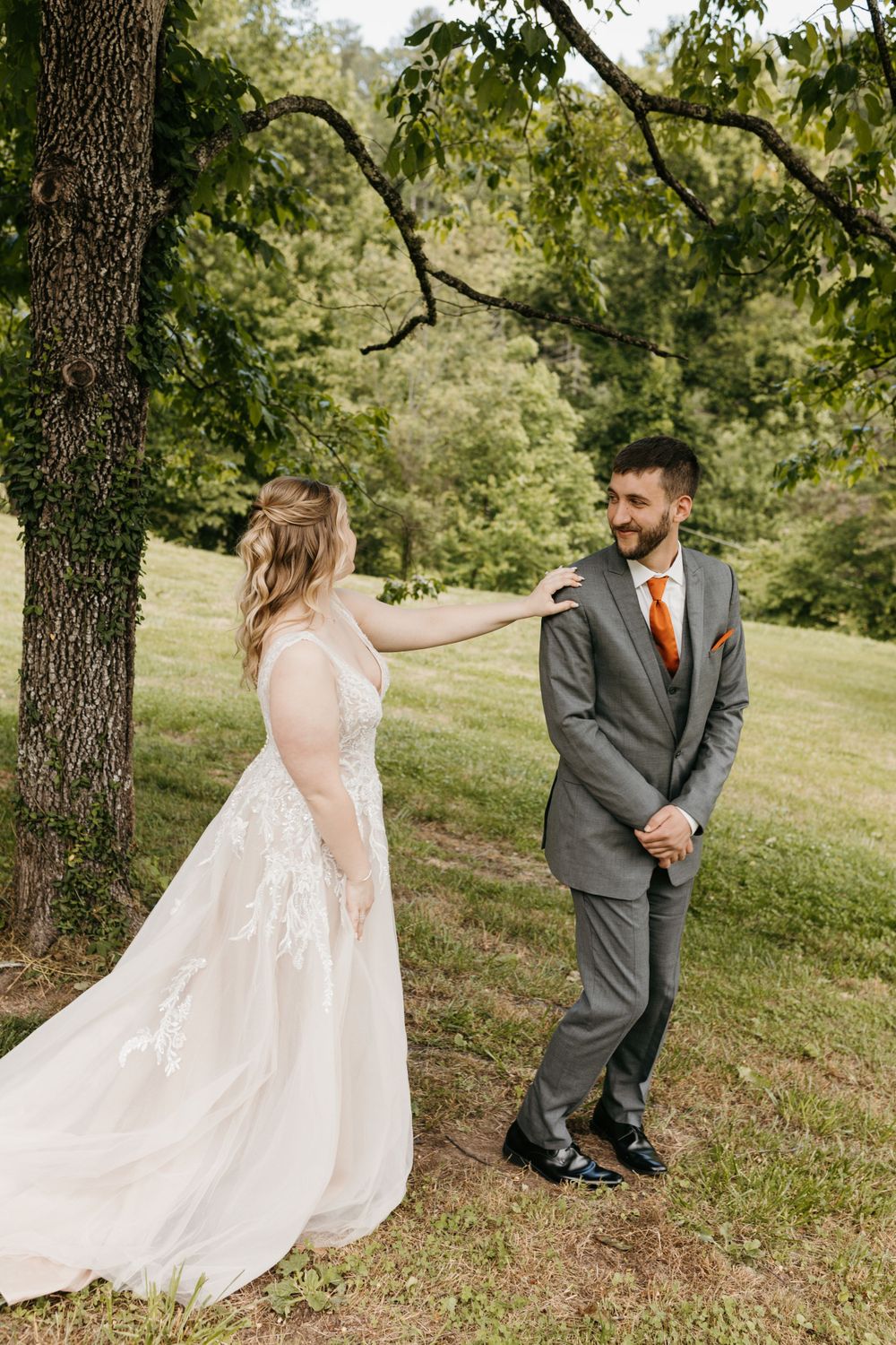 Groom in gray suit with orange tie smiles as bride in lace wedding gown approaches him for first look.