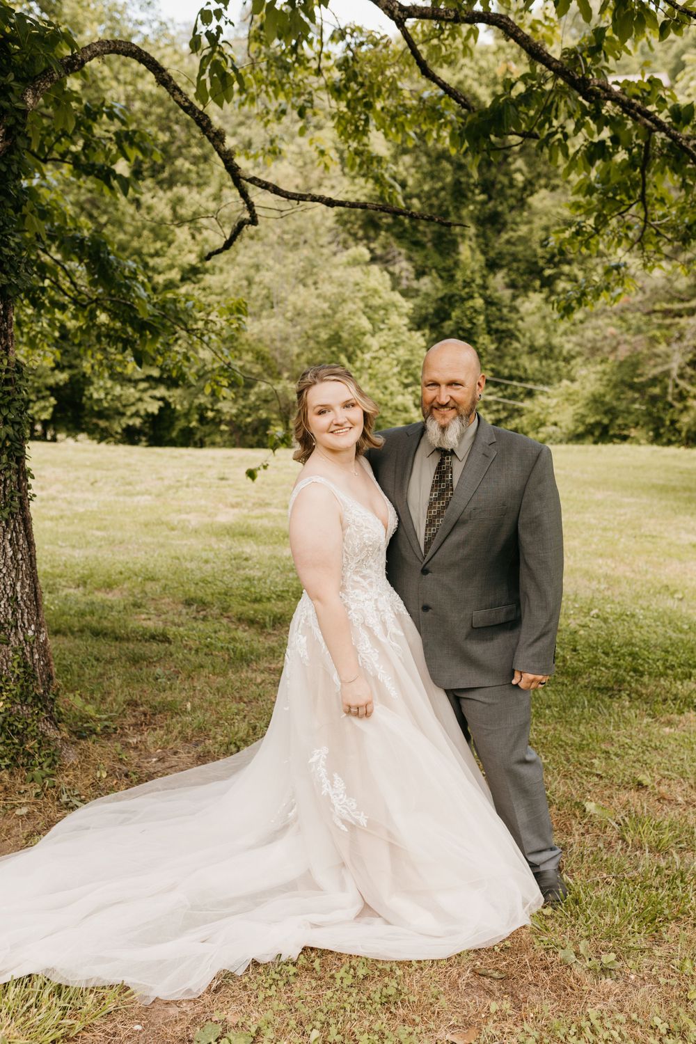Bride in a flowing lace gown and bearded groom in gray suit pose together under a tree in a lush green field.