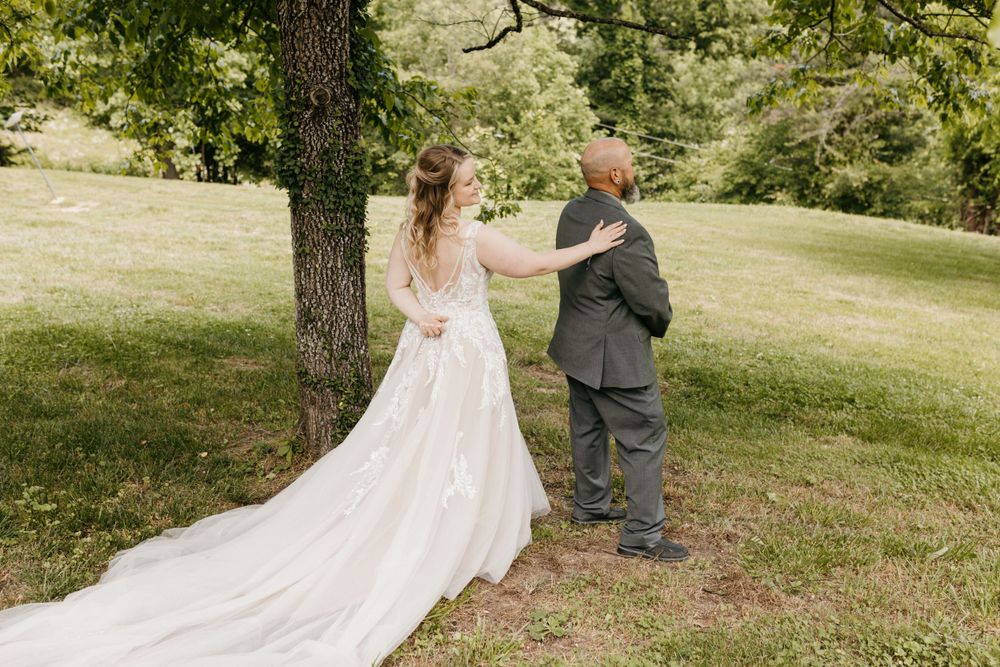 Bride surprising groom during first look under a tree in a lush green outdoor setting.