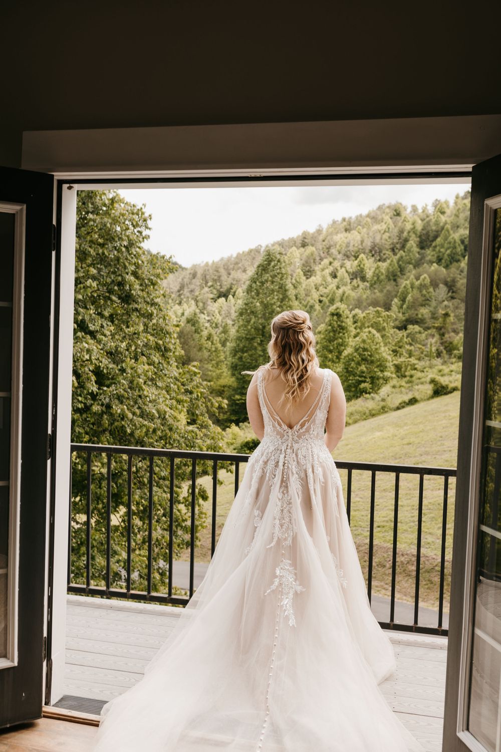 Bride in lace ballgown standing on balcony overlooking green forested mountain landscape.