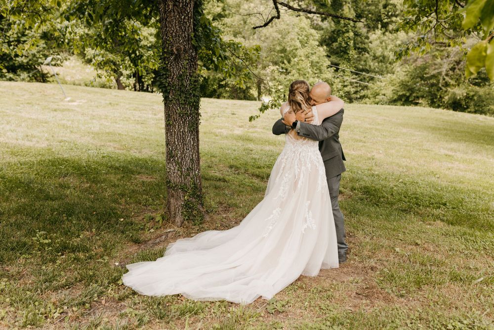Bride and groom share a tender embrace under a large tree on a green hillside during their outdoor wedding.