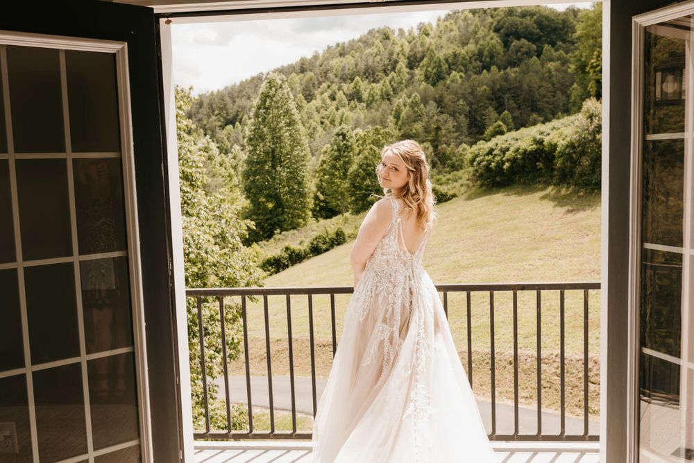 Bride in elegant lace wedding gown turning on a balcony with scenic green hills behind her.