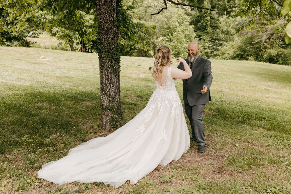 Groom turning to see bride in flowing lace gown during outdoor first look under large tree.