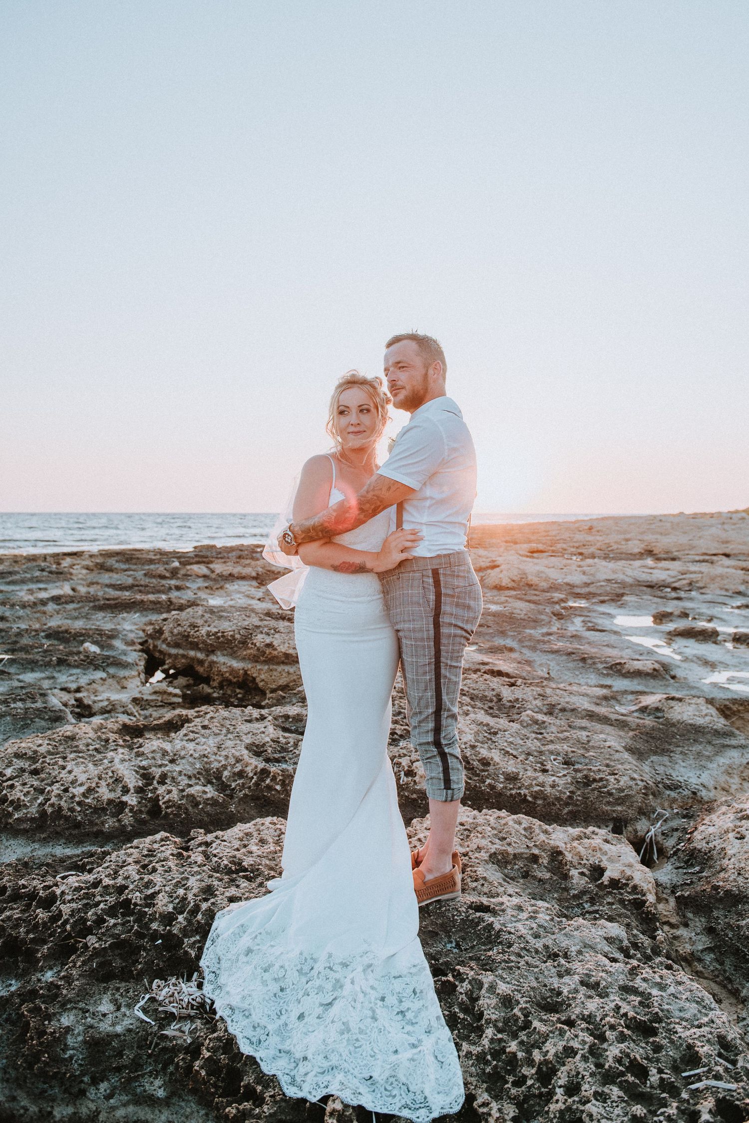 A romantic seaside wedding portrait at sunset on rocky coastal terrain.