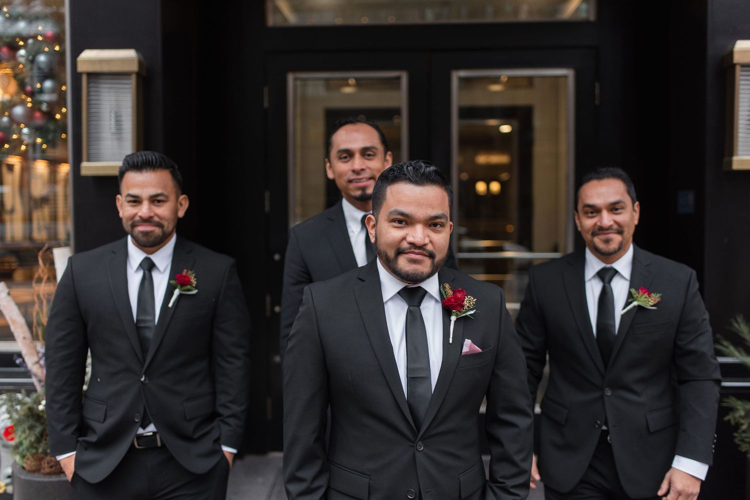 A group of groomsmen in matching dark suits pose outside a building during a wedding celebration.