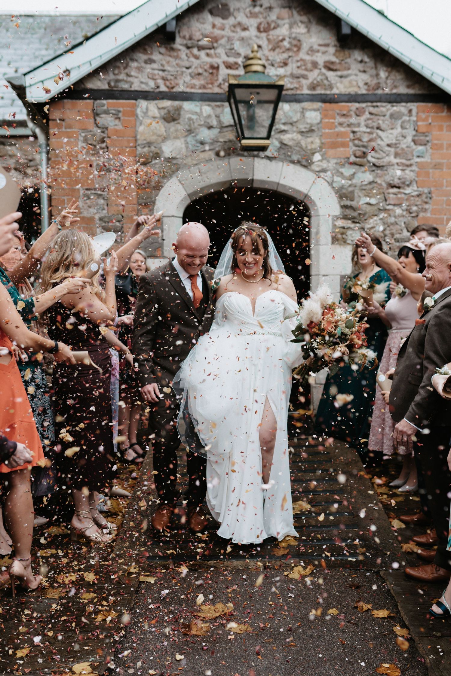 Wedding celebration with guests throwing confetti at couple exiting stone building.