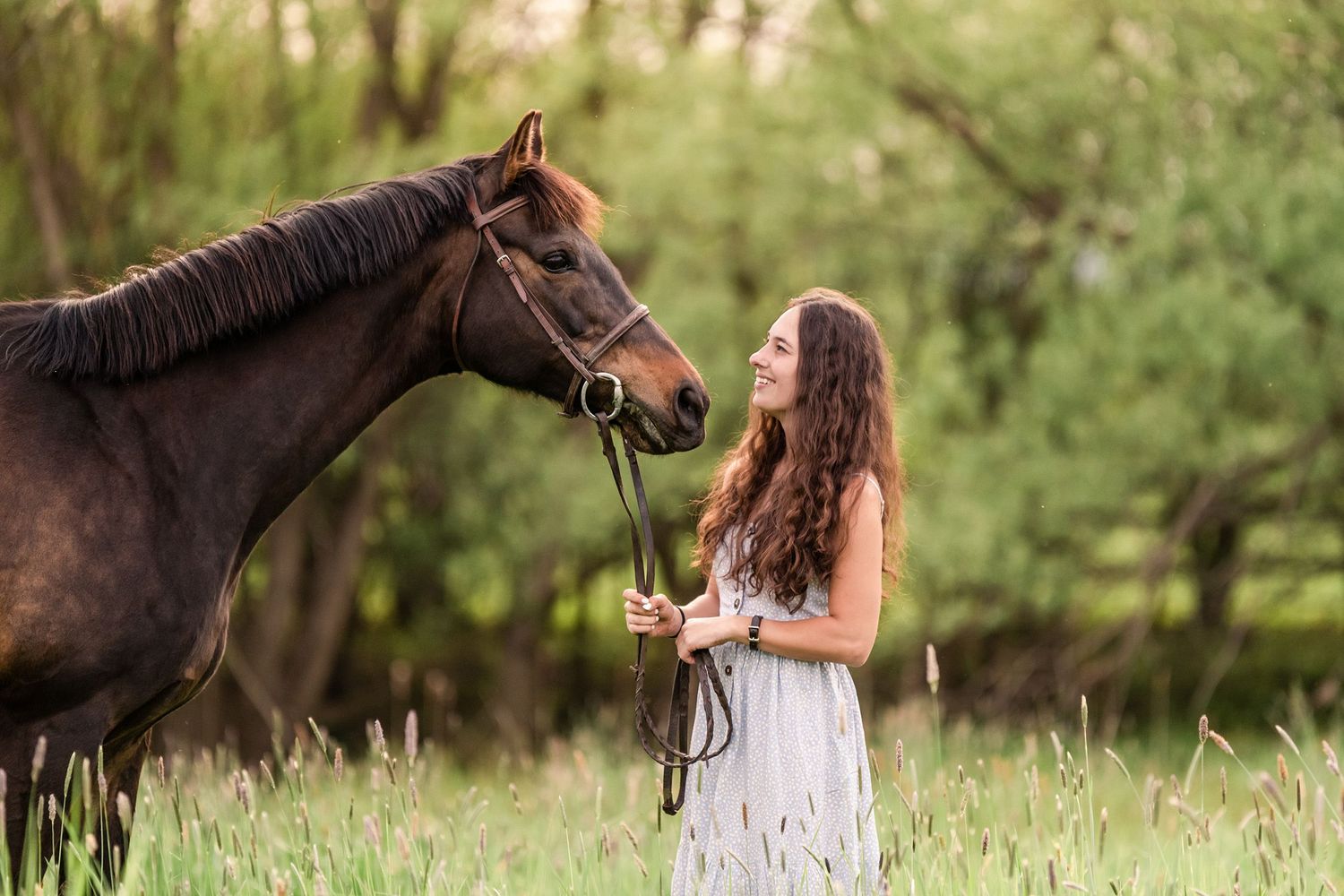 Girl in a pale blue dress smiling and staring into the eyes of a bay horse