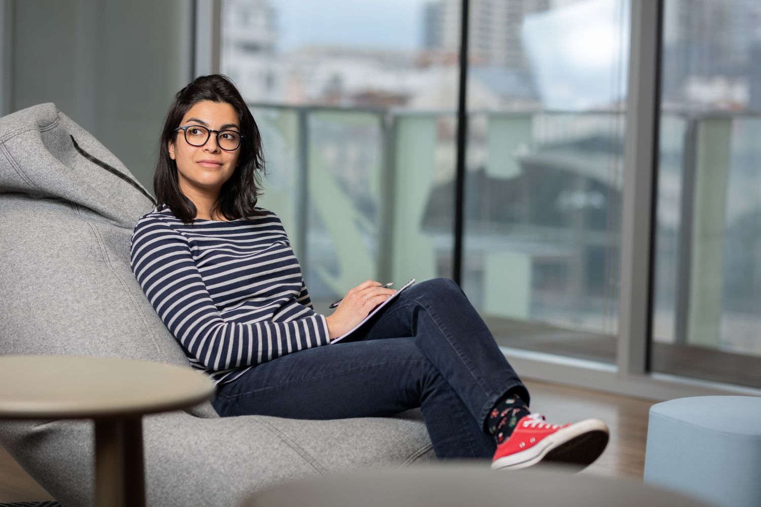 Person in glasses wearing striped shirt relaxes on grey chair by window in modern space.
