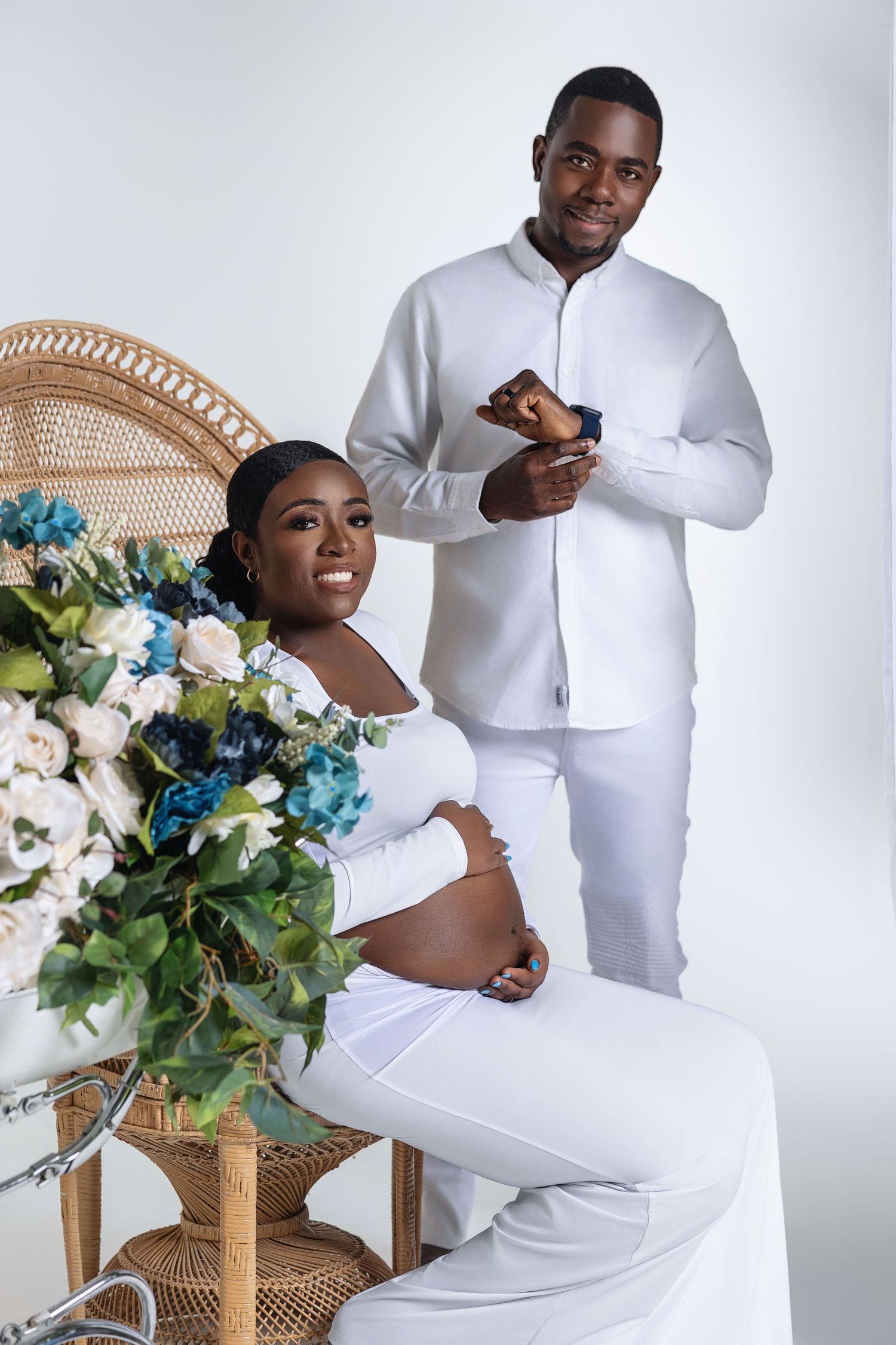Expecting couple in white outfits posing with floral arrangement and wicker chair for maternity shoot.