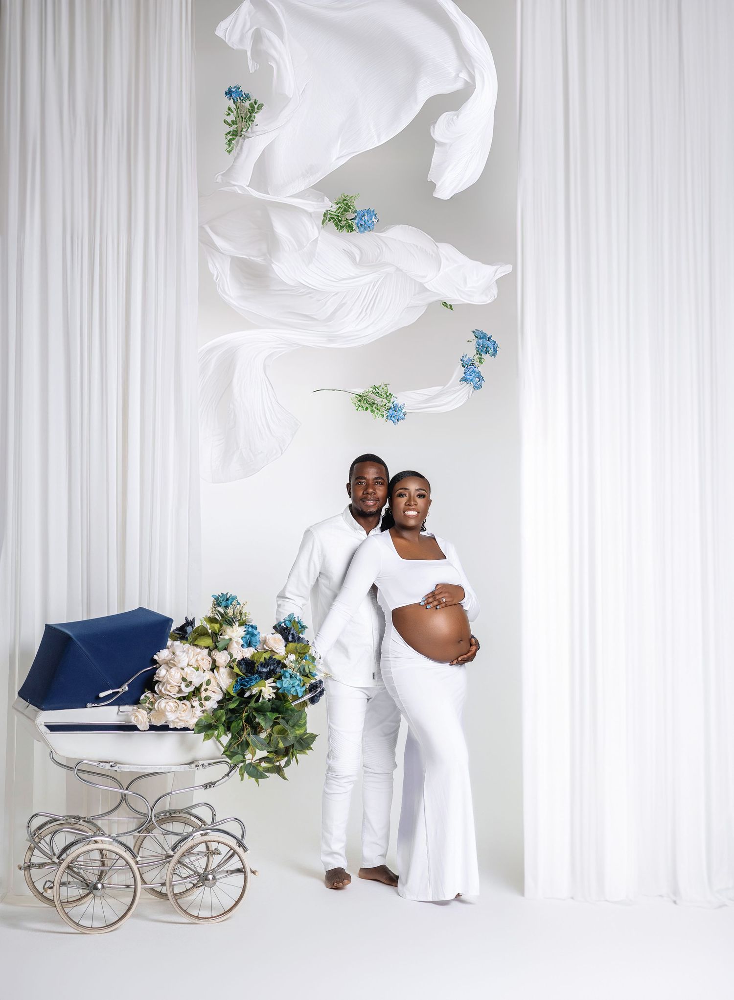 Couple in white maternity photoshoot with flowing fabric and blue floral vintage baby carriage backdrop.