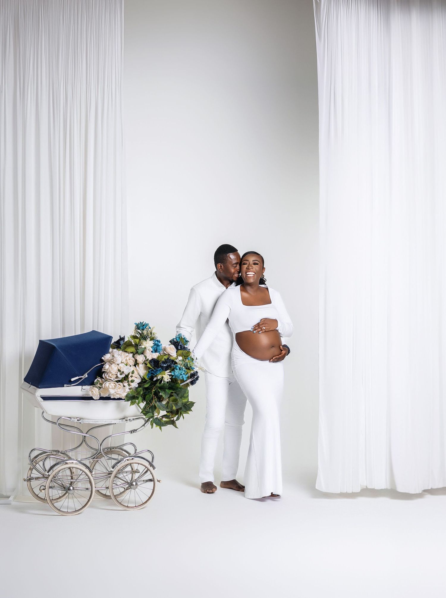 Elegant maternity photoshoot with couple in white outfits beside a vintage baby carriage with blue and white flowers.