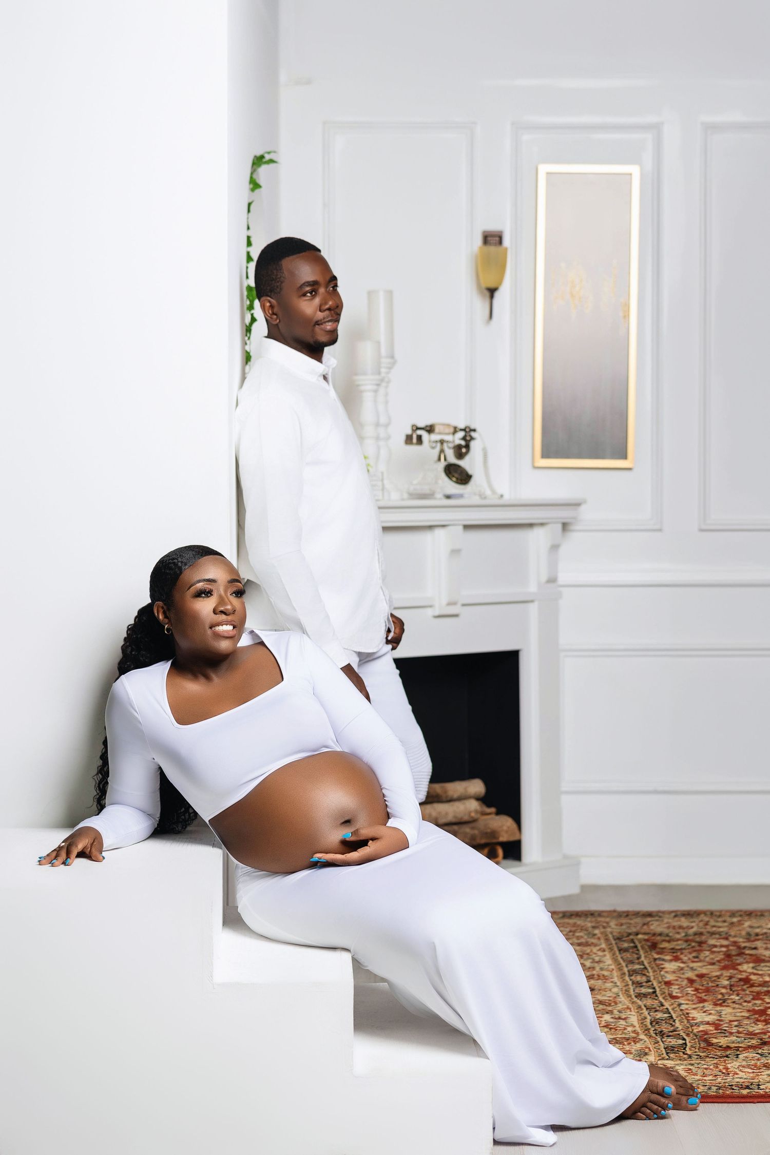 Pregnant woman in white outfit sits on steps while partner stands behind her in a bright white room.
