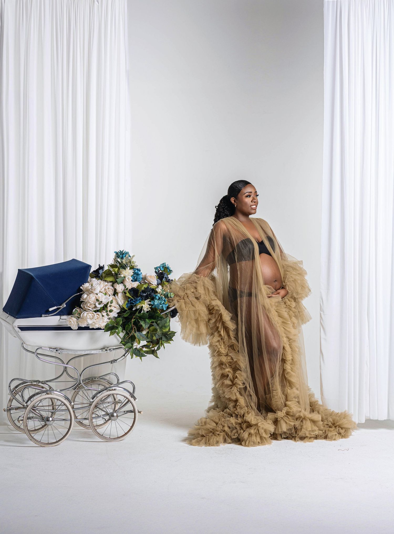 Pregnant woman in gold tulle robe poses beside a vintage navy baby carriage with floral arrangement.