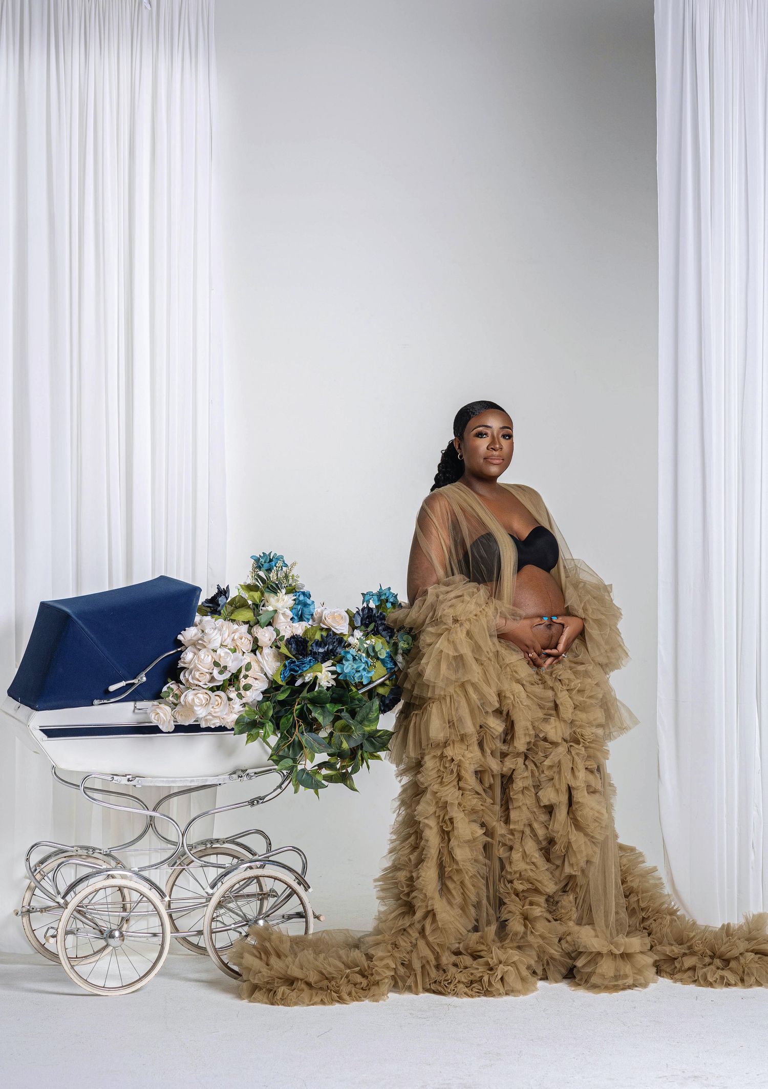 Expectant mother in gold ruffled tulle gown stands next to a vintage baby carriage with blue flowers.