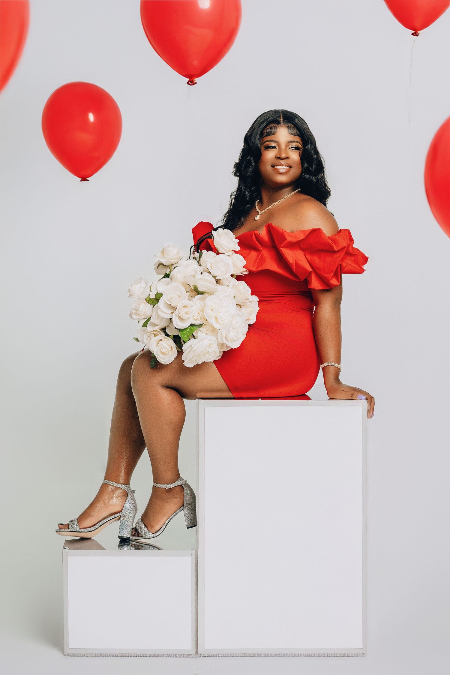 Woman in red off-shoulder dress holding white roses seated on white cube with red balloons.